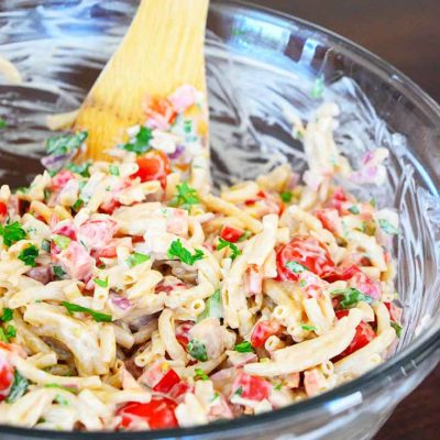 A glass bowl sits filled with just-mixed, clean eating easy macaroni salad. A wooden spoon is nestled into the salad at the side of the bowl.