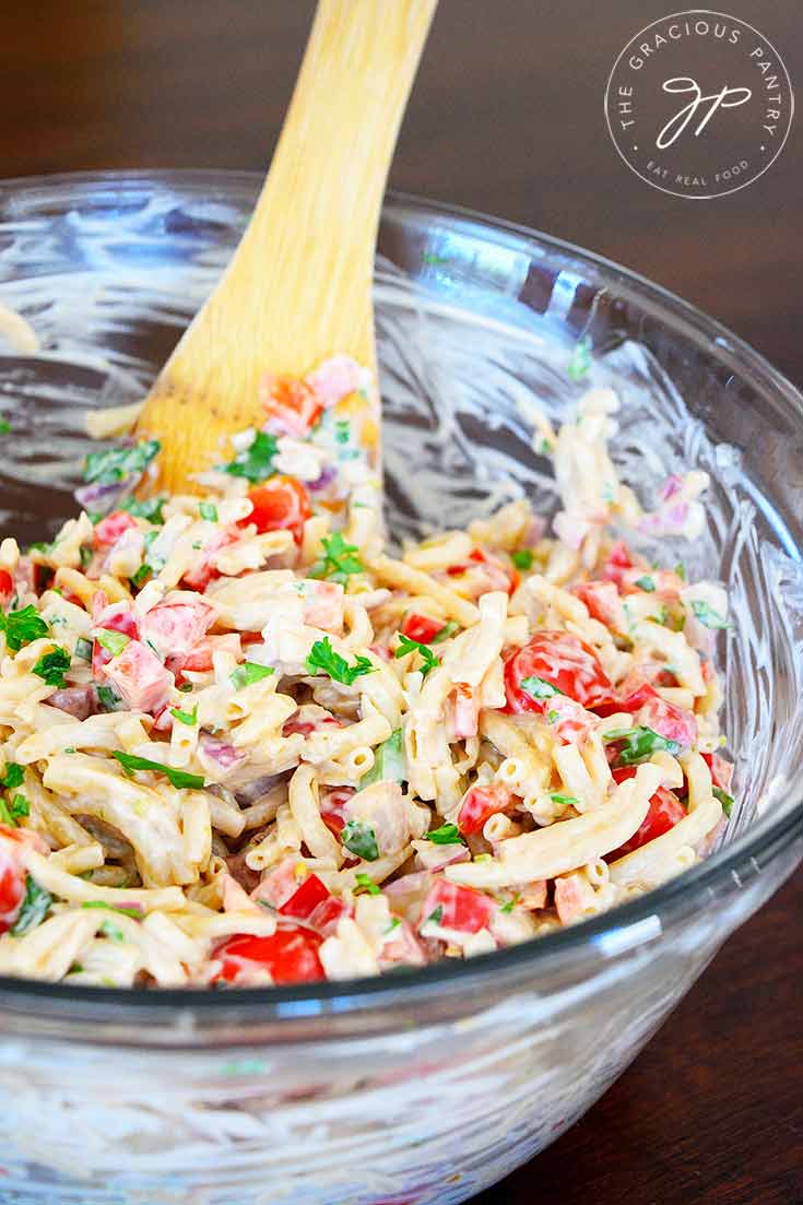 A glass bowl sits filled with just-mixed, clean eating easy macaroni salad. A wooden spoon is nestled into the salad at the side of the bowl.