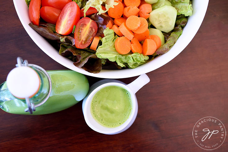 And overhead view looking down into the bowl of salad with carrots, tomatoes and cucumbers. A clear glass bottle and small white pitcher both sit in front of the bowl filled with this clean eating green goddess dressing recipe.