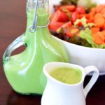 A beautiful bowl of salad sits towards the back of the table with a full dressing bottle and a small, white pitcher sit filled with this clean eating green goddess dressing recipe.