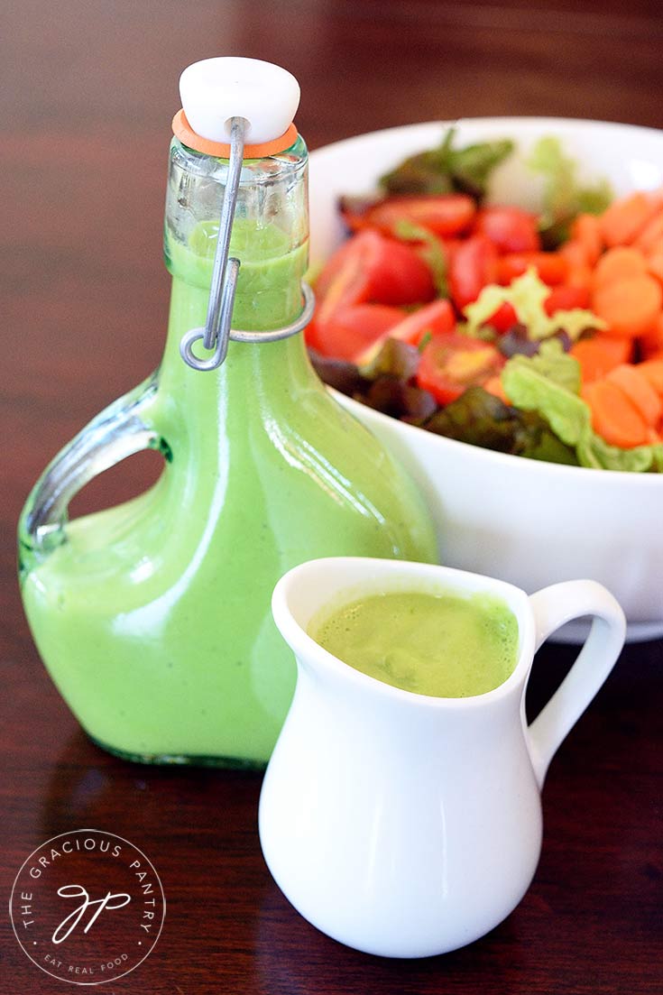 A beautiful bowl of salad sits towards the back of the table with a full dressing bottle and a small, white pitcher sit filled with this clean eating green goddess dressing recipe.