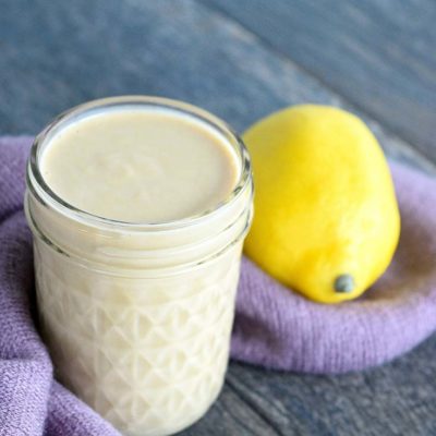 A single jar of this clean eating lemon tahini dressing recipe sits on a gray surface with a purple towel at it's base and a lemon to the right.
