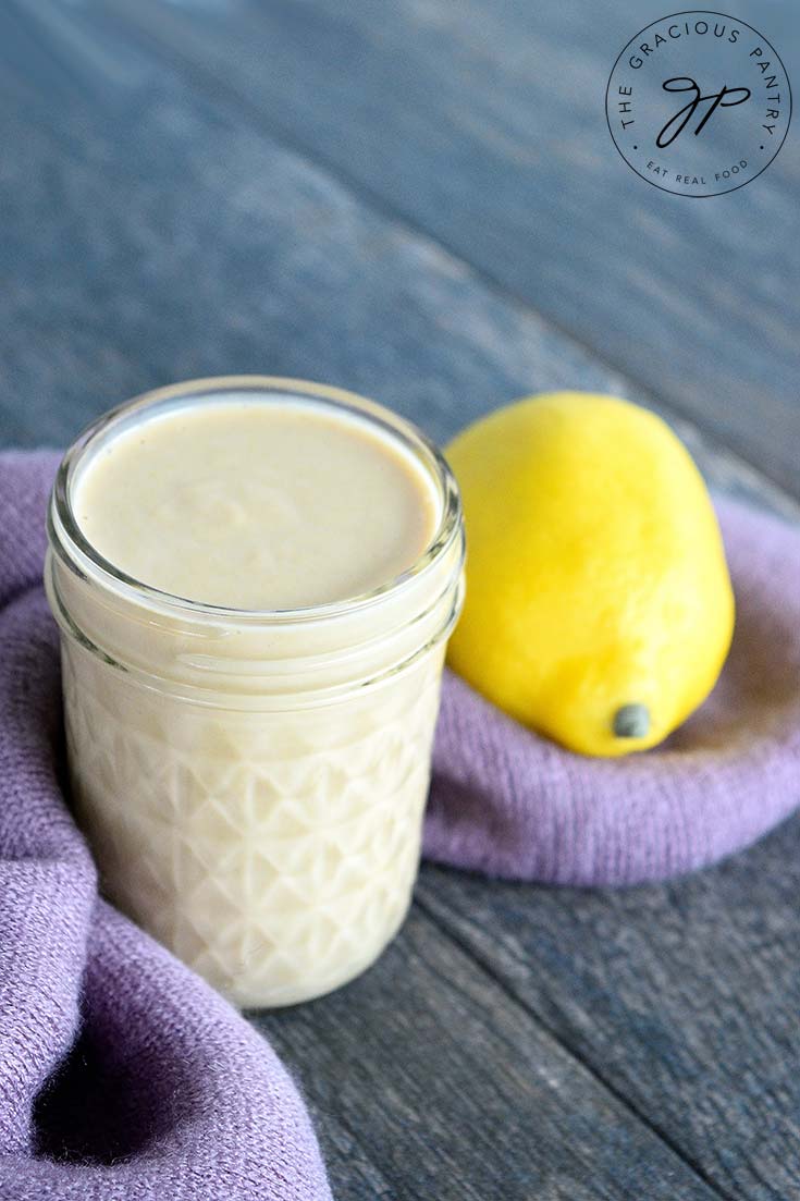 A single jar of this clean eating lemon tahini dressing recipe sits on a gray surface with a purple towel at it's base and a lemon to the right.