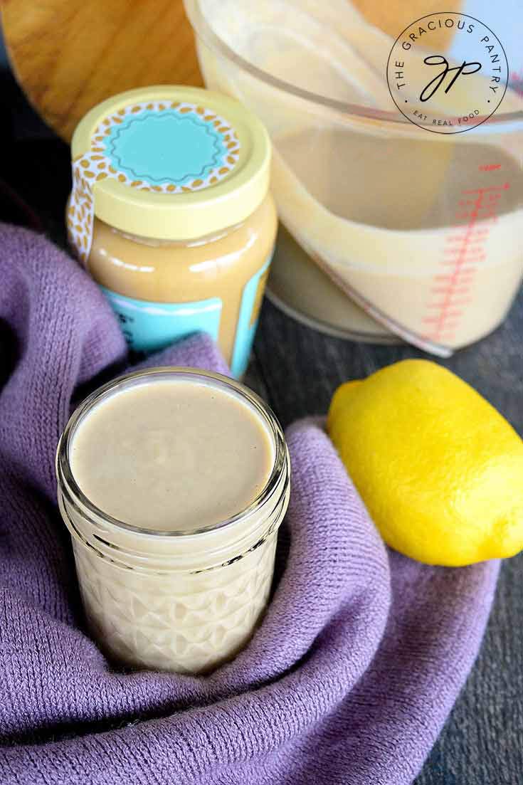 An open canning jar filled with this clean eating lemon tahini dressing sits next to an unopened jar of tahainin, a measuring cup that still contains some of the dressing and a lemon.