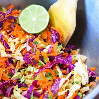 This fresh, clean eating Mexican coleslaw recipe is being served in a silver, metalic bowl. It has a half of a lime wedged in at the side of the bowl and a wooden serving spoon sits nested at the side of the bowl as well.