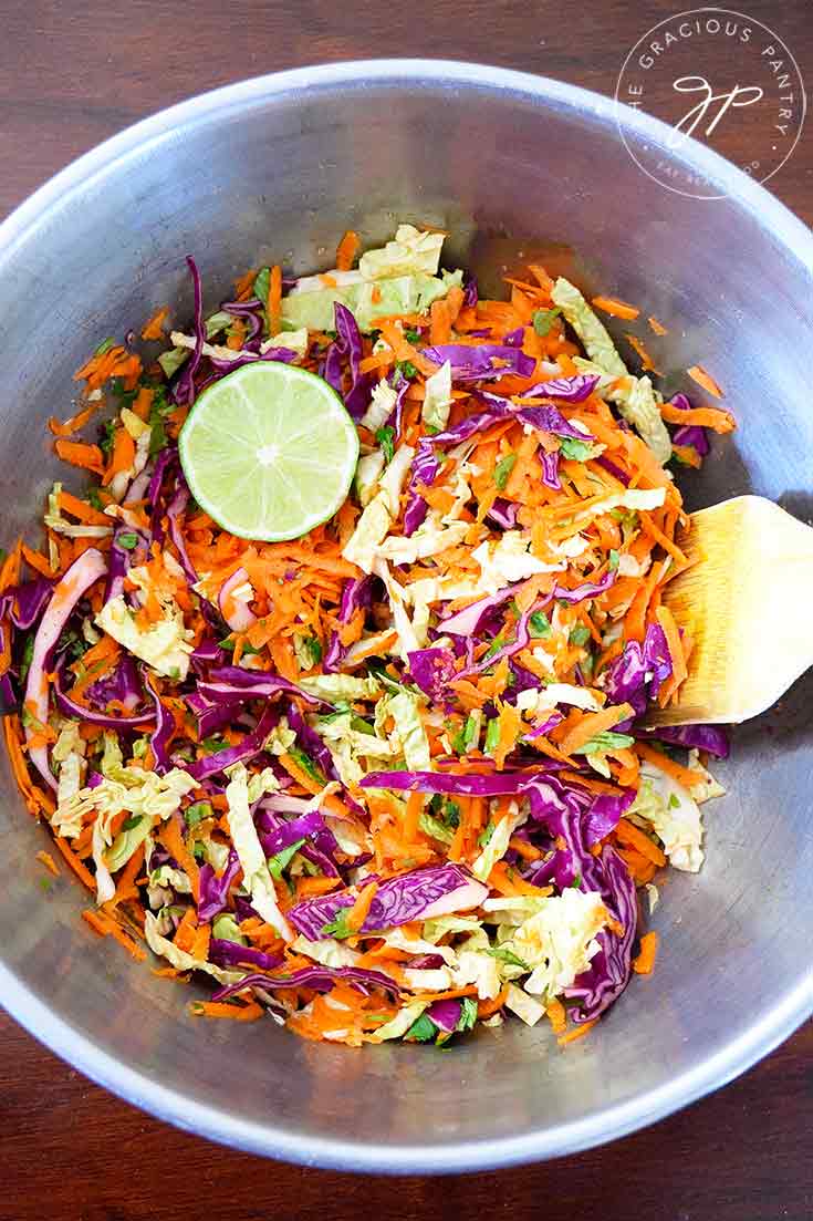 An overhead view looking down into a metalic bowl at this clean eating Mexican coleslaw recipe. You can see half a lime, a wooden serving spoon and the brightly colored green, purple and orange coleslaw.