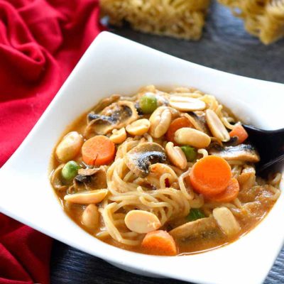 A white bowl filled with this clean eating Thai ramen soup recipe sits with a spoon in the bowl, ready to eat. Two dry blocks of ramen sit behind the bowl and a red napkin sits to the left of the bowl.