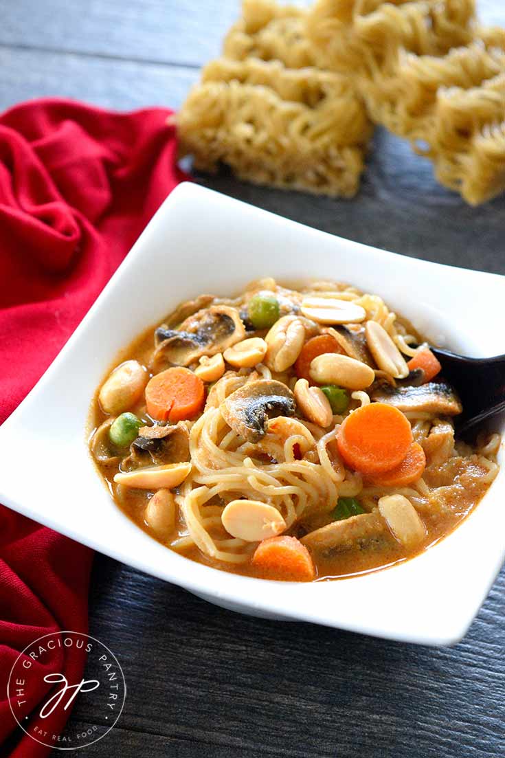 A white bowl filled with this clean eating Thai ramen soup recipe sits with a spoon in the bowl, ready to eat. Two dry blocks of ramen sit behind the bowl and a red napkin sits to the left of the bowl.