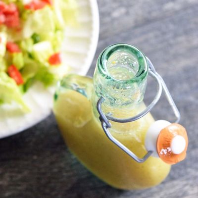 An overhead view of a bottle of this Mustard Vinaigrette sitting next to a green salad with red tomatoes, ready to eat.