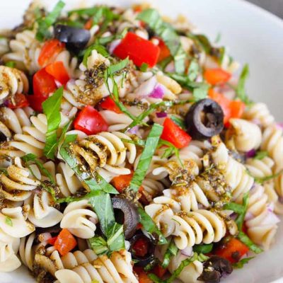 A white bowl sits filled with this Clean Eating Rustic Italian Pasta Salad Recipe. You can see bits of olives, red peppers, fresh basil and a bit of the dressing scattered throughout the pasta.