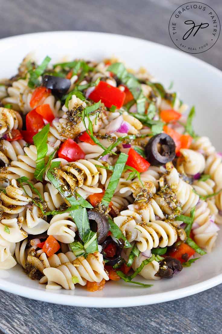 A white bowl sits filled with this Clean Eating Rustic Italian Pasta Salad Recipe. You can see bits of olives, red peppers, fresh basil and a bit of the dressing scattered throughout the pasta.