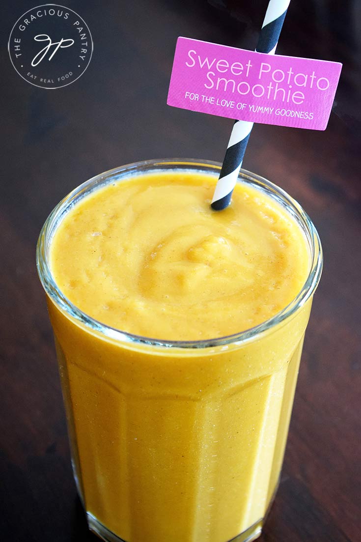 A closeup shot of this clean eating sweet potato smoothie shows the golden colored drink in a clear, tall glass with a black and white stripped straw.