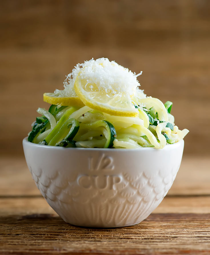 A white bowl sits filled with this Clean Eating Lemon Parmesan Zucchini Noodles on a wooden table. A lemon slice sits on top of the zucchini noodles and everything is topped with freshly grated parmesan cheese.