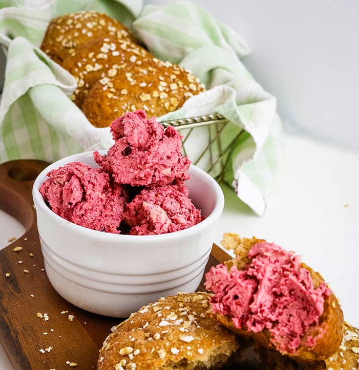 A white bowl filled with this Triple Berry Honey Butter sits next to bread rolls and a single roll is cut and topped with this delicious spread