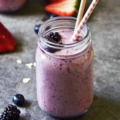 A head on view of a mason jar filled with this Mixed Berry Smoothie. It has two straws in it and a couple of blackberries. Several cut strawberries sit in the background.