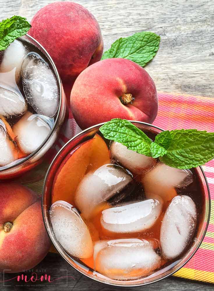 An overhead view of this clean eating peach iced tea recipe shows two glasses sitting on a table next to a peach, filled with the dark brown tea, ice cubes and some fresh mint.