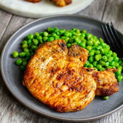 A plate of air fried pork chops sits next to a side of peas with left over pork chops sitting on a board to the side.