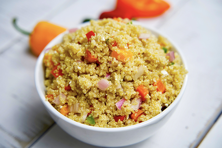 A side view of a white bowl filled with this multi-colored, Clean Eating Mediterranean Quinoa Salad. A red and orange pepper lay on the table just behind the bowl.