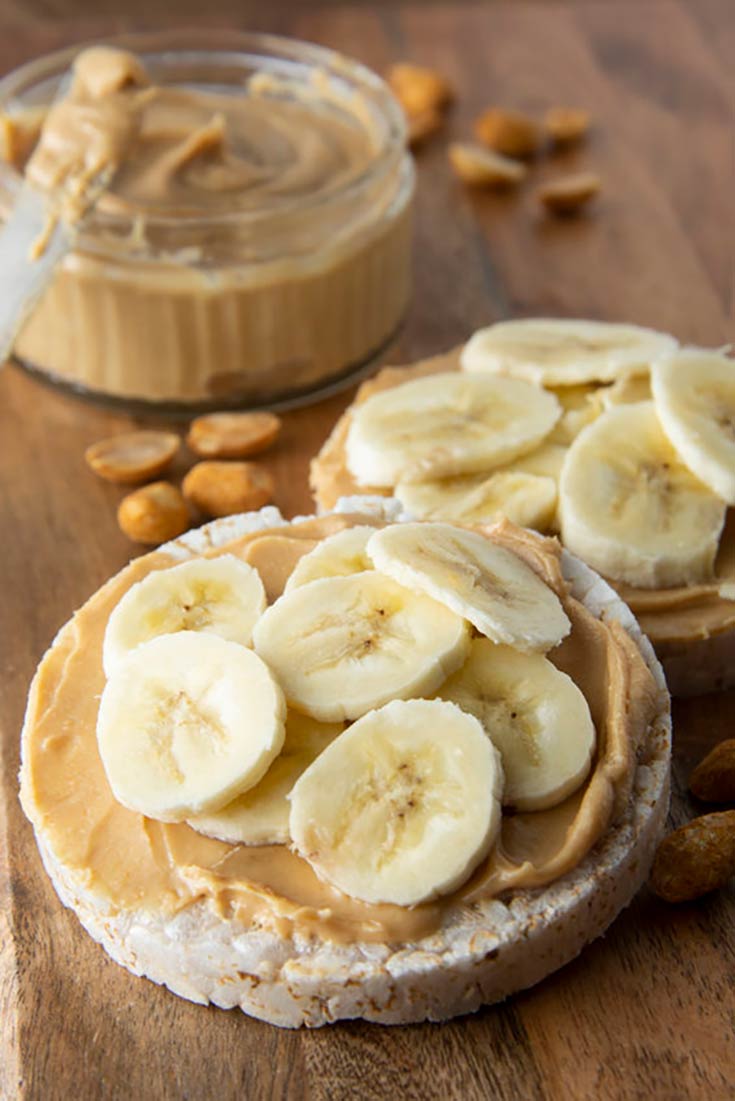 Two rice crackers sit on a cutting board with peanut butter spread on and banana slice piled on top in this guide to After School Snacks That Won't Ruin Dinner