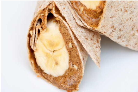 A cut banana wrap sits on a white background. You can see the layers of the whole grain wrap, nut butter and the bananas.