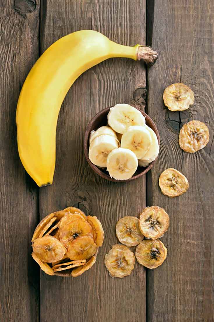 A single, whole banana lays on a wooden table top with a dish of sliced bananas next to it as well as dried banana chips strewn about.