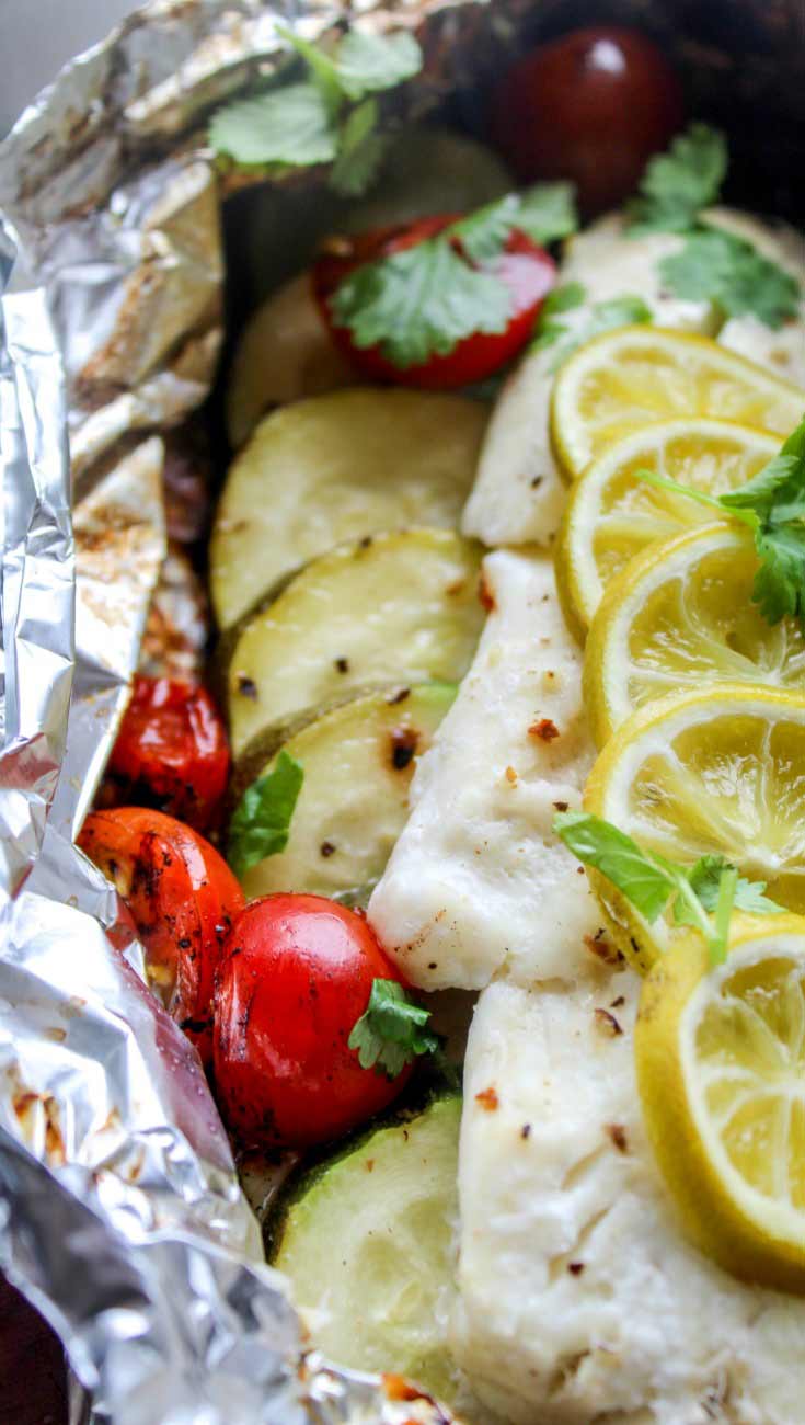 A white fish fillet sitting on a bed of sliced zucchini with some red cherry tomatoes tucked in the sides. The fish has lemon slices over the top.