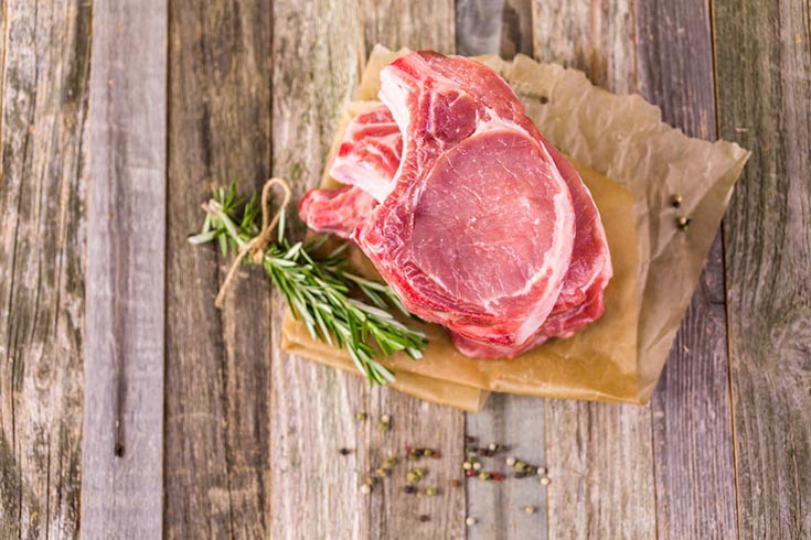 An overhead view of two pork chops stacked on top of each other with a sprig of fresh rosemary sitting next to them.