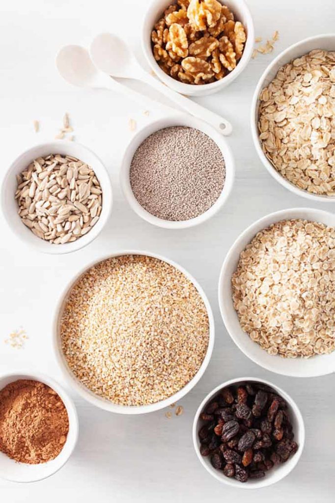 Bowls of different types of oats and oatmeal toppings sit on a white background.