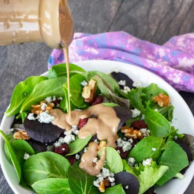A beautiful bowl of cranberry walnut salad sits on a table and this blue cheese vinaigrette is being poured over the top.