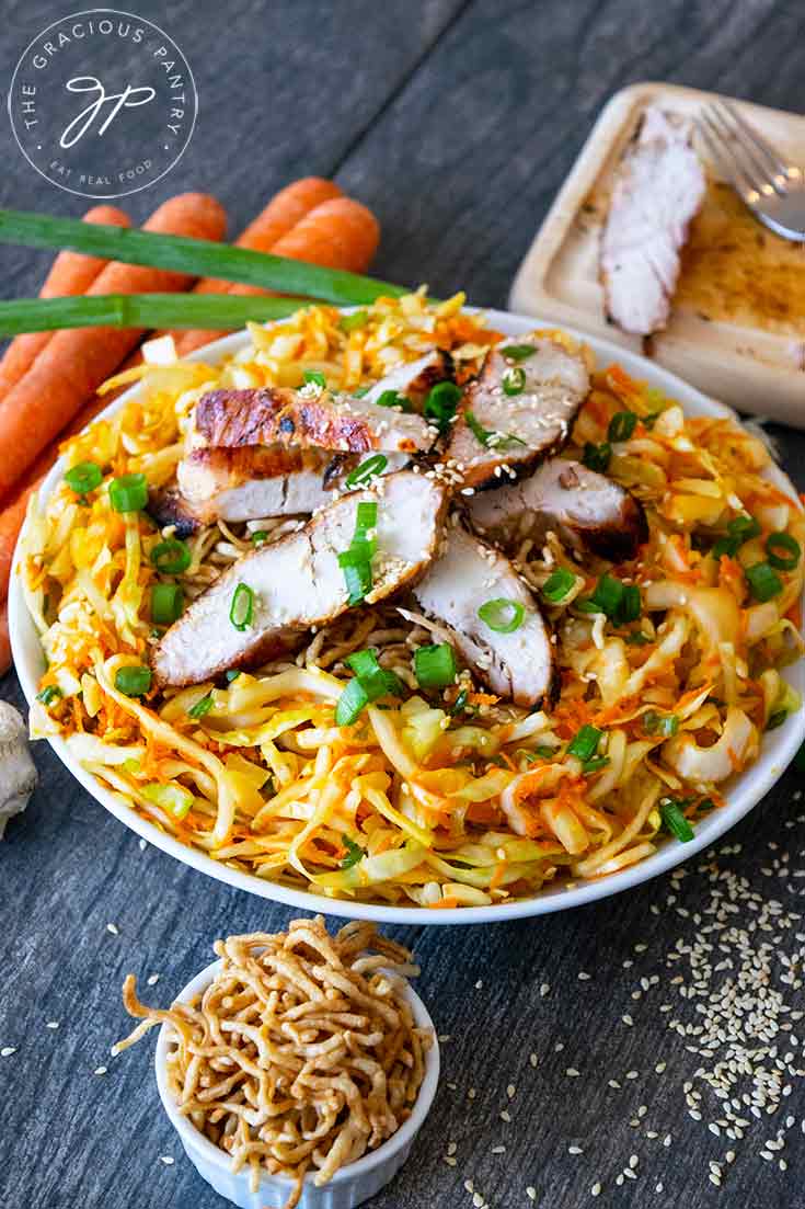 This Chinese Chicken Salad sits surrounded by carrots, green onions, ramen noodles and a small cutting board with a fork where the chicken was sliced.