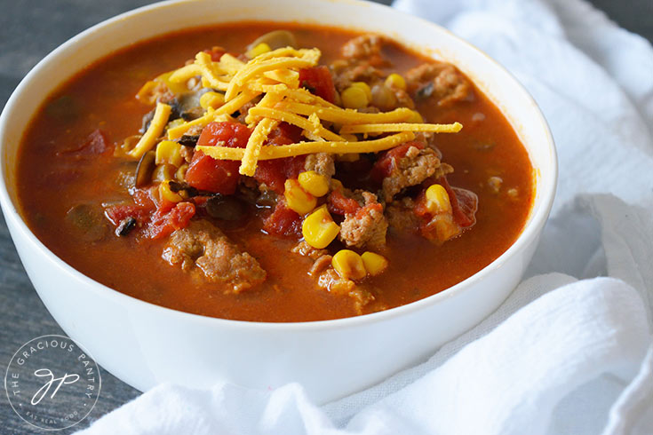 A white bowl of chili with a white napkin next to it. A brown broth surrounds the meat, corn, tomatoes and shredded cheese at the center.