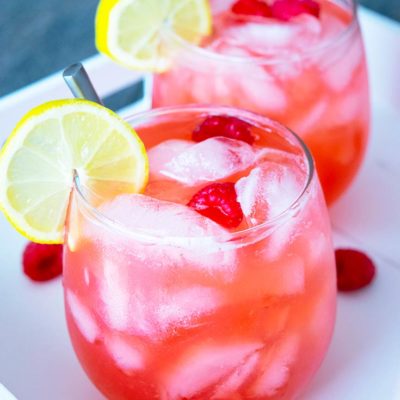 Two freshly poured glasses of raspberry lemonade sit on a serving tray, ready to drink. They each have a lemon slice on the side of the glass and two raspberries floating on top.