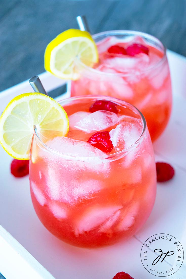 Two freshly poured glasses of raspberry lemonade sit on a serving tray, ready to drink. They each have a lemon slice on the side of the glass and two raspberries floating on top.