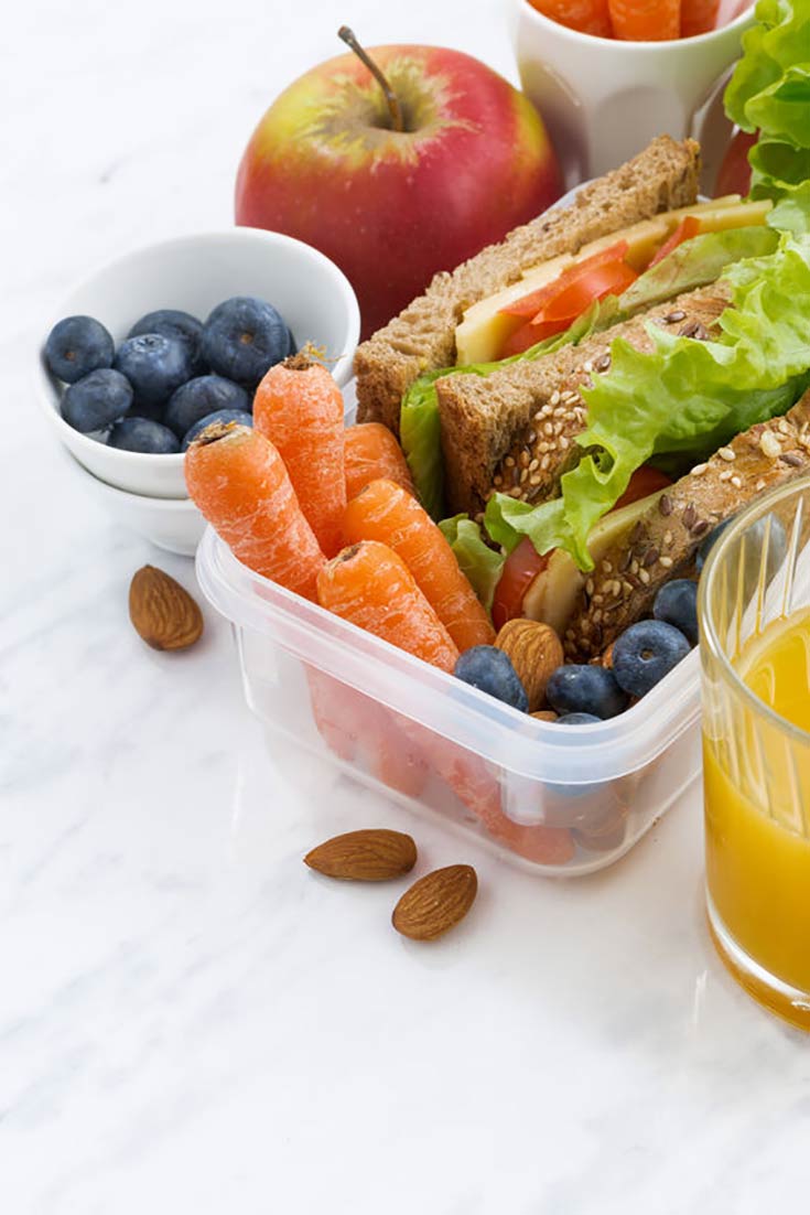 A healthy school lunch sits ready to eat. There are blueberries, carrots, an apple and a whole grain sandwich in lunch containers.