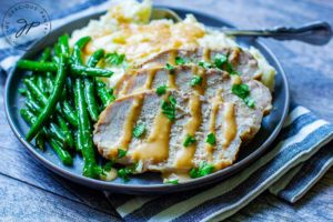 The last step of this Slow Cooker Pork Loin Recipe shows the pork sliced and sitting on a plate next to mashed potatoes and green beans, with gravy drizzled over the top of the meat and potatoes.