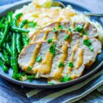 Three slices of this Slow Cooker Pork Loin is sliced and placed on a plate next to mashed potatoes and green beans. Gravy dresses the pork and potatoes.