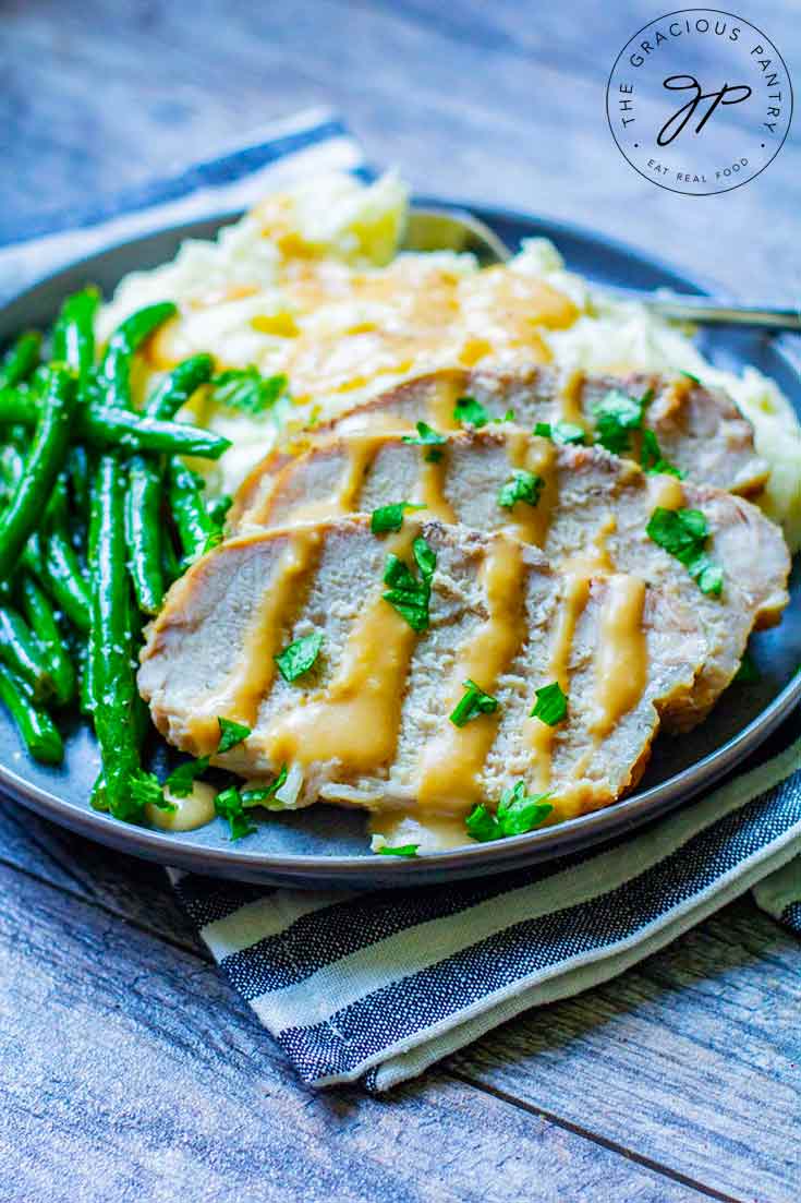 Three slices of this Slow Cooker Pork Loin is sliced and placed on a plate next to mashed potatoes and green beans. Gravy dresses the pork and potatoes.