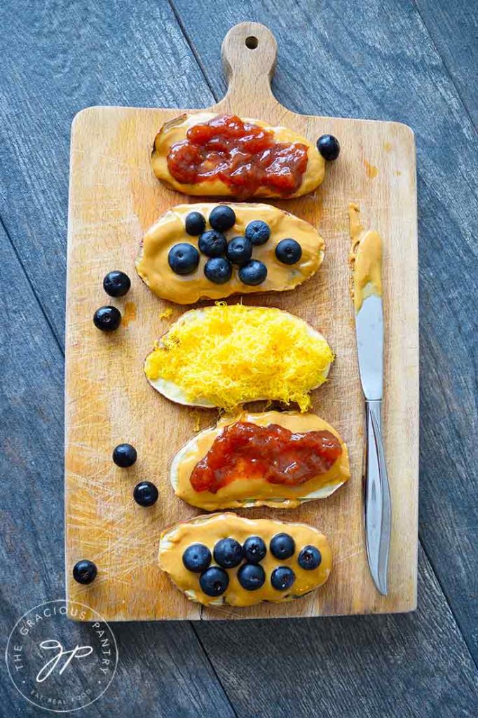 A line of sweet potato slices sit on a cutting board, topped with various toppings such as, peanut butter and jelly, peanut butter and blueberries and the one in the middle is topped with grated cheddar cheese.