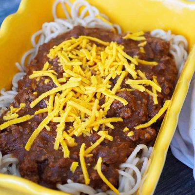 A close up view of a yellow casserole dish containing some pasta on the bottom level, Cincinnati chili in the middle, ad some grated cheddar cheese on top.