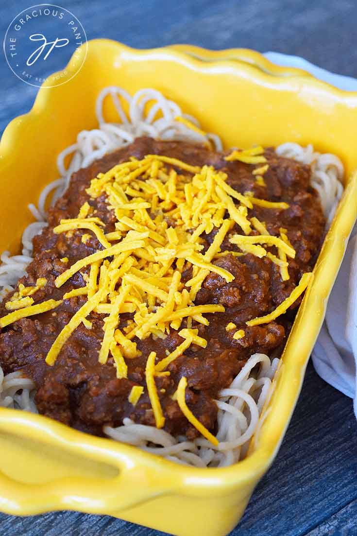 A close up view of a yellow casserole dish containing some pasta on the bottom level, Cincinnati chili in the middle, ad some grated cheddar cheese on top.