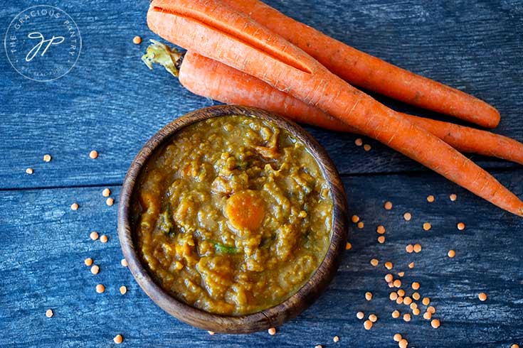 An overhead view of this sweet potato curry looks down into a wooden bowl filled with the curry. Whole carrots sit to the upper right side of the bowl and dry lentils are strewn around the bowl on the table.