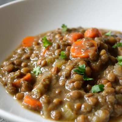 A white bowl sits on a table with a nice helping of this Vegan Instant Pot Lentil Soup. You can see carrot slices in with the lentils and fresh herbs sprinkled over the top.