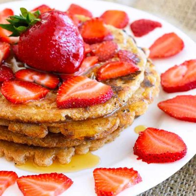 A stack of spelt pancakes sits on a white plate, topped with fresh strawberries and maple syrup.