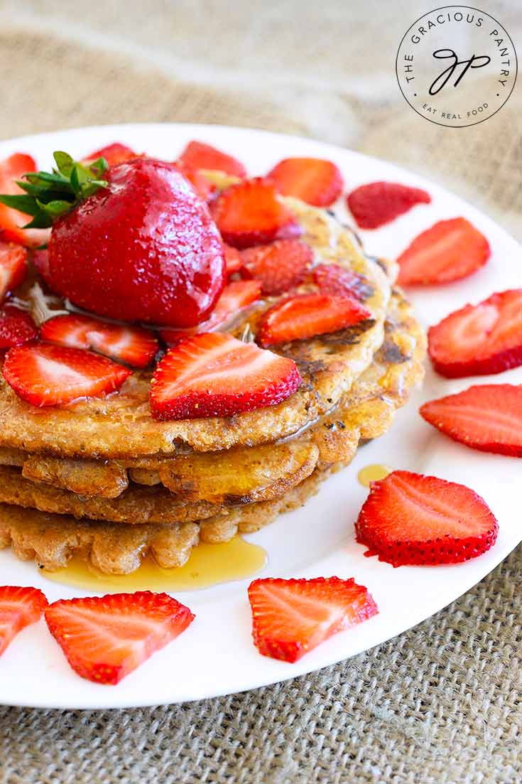 A stack of spelt pancakes sits on a white plate, topped with fresh strawberries and maple syrup.