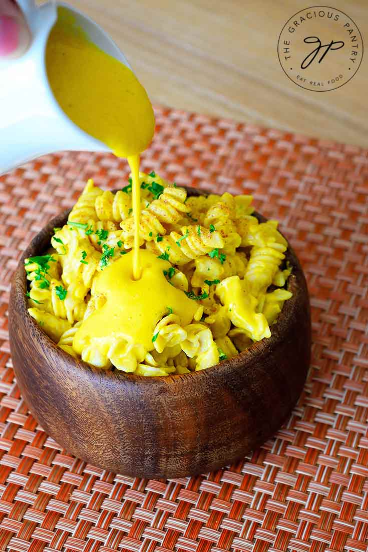 A wooden bowl of pasta sits on a table and a pitcher of this Butternut Squash Pasta Sauce is being poured over the top of it.