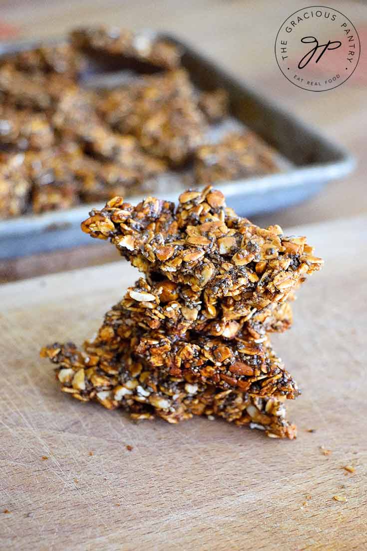 A stack of thisPumpkin Spice Granola Bark sits on a cutting board with the baking pan sitting behind it.