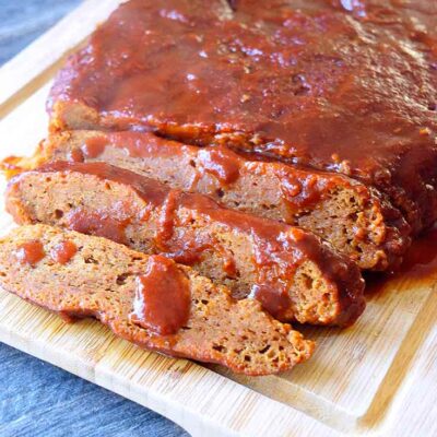 A Seitan Brisket With Barbecue Glaze sits partially sliced on a cutting board with barbecue sauce drizzled over the top of the brisket.