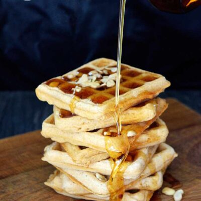 A stack of waffles made from this Vegan Waffles Recipe, are having maple syrup poured over the top. It's drizzling down the side into an amber pool of syrup at the bottom.