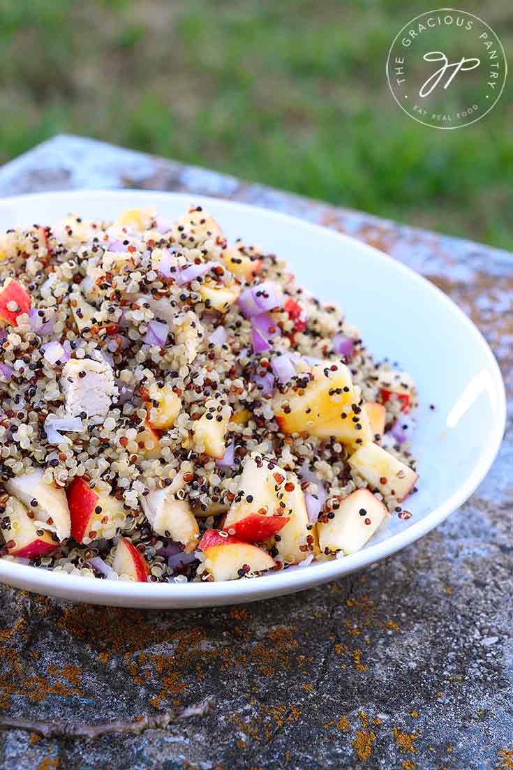 A bowl of this Apple Quinoa Salad With Chicken sits on a stone bench with grass in the background. You can see the quinoa, apples and chunks of chicken.