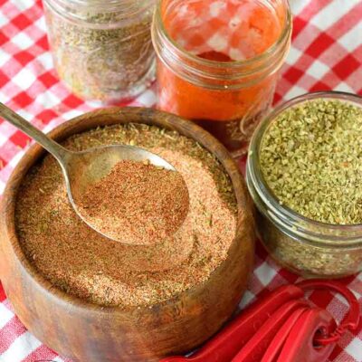A wooden bowl filled with this Cajun Seasoning Recipe, sits with a serving spoon in the bowl and jars of spices and measuring spoons around it.